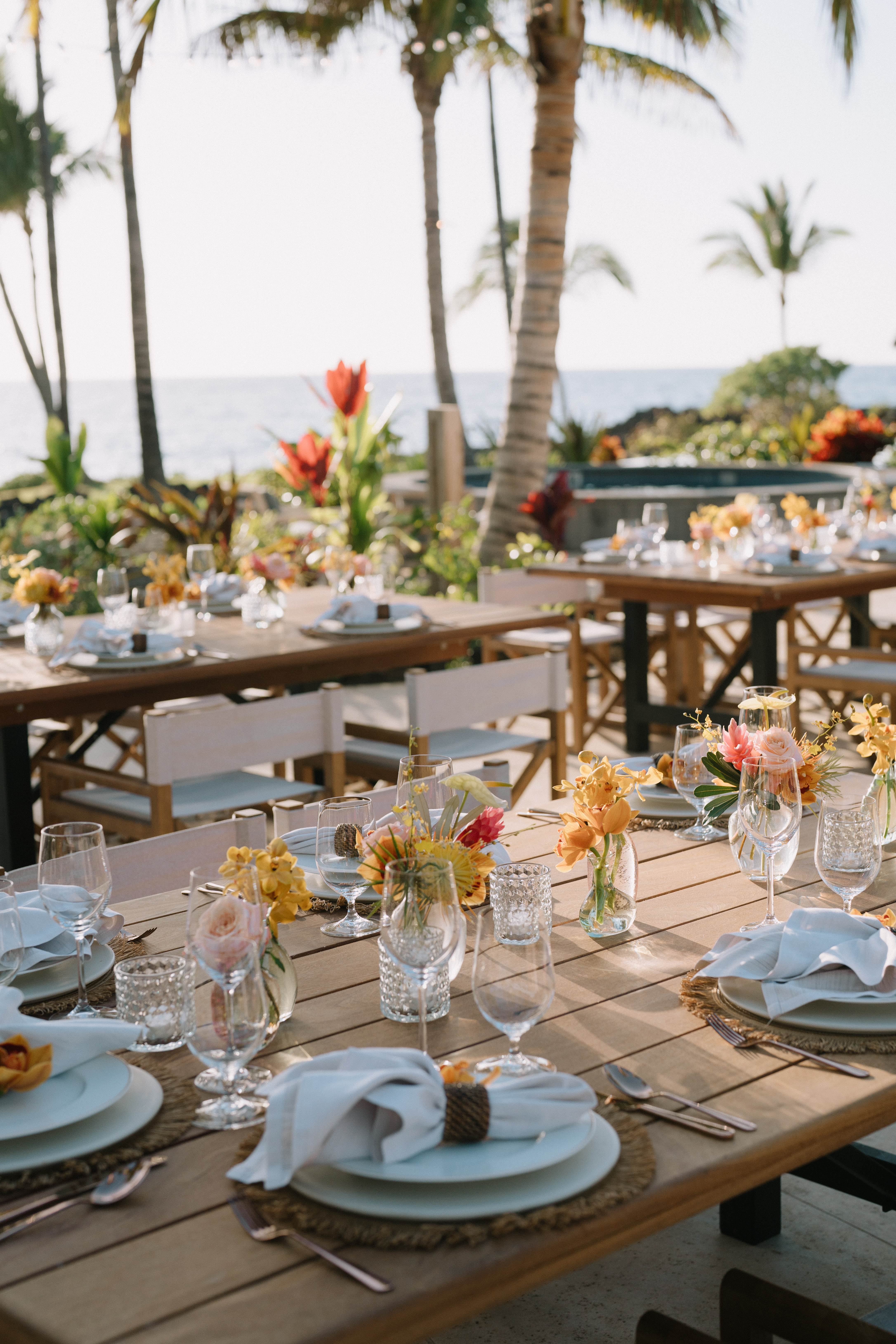 Elegant outdoor table setup with floral centerpieces, overlooking ocean and palm trees.