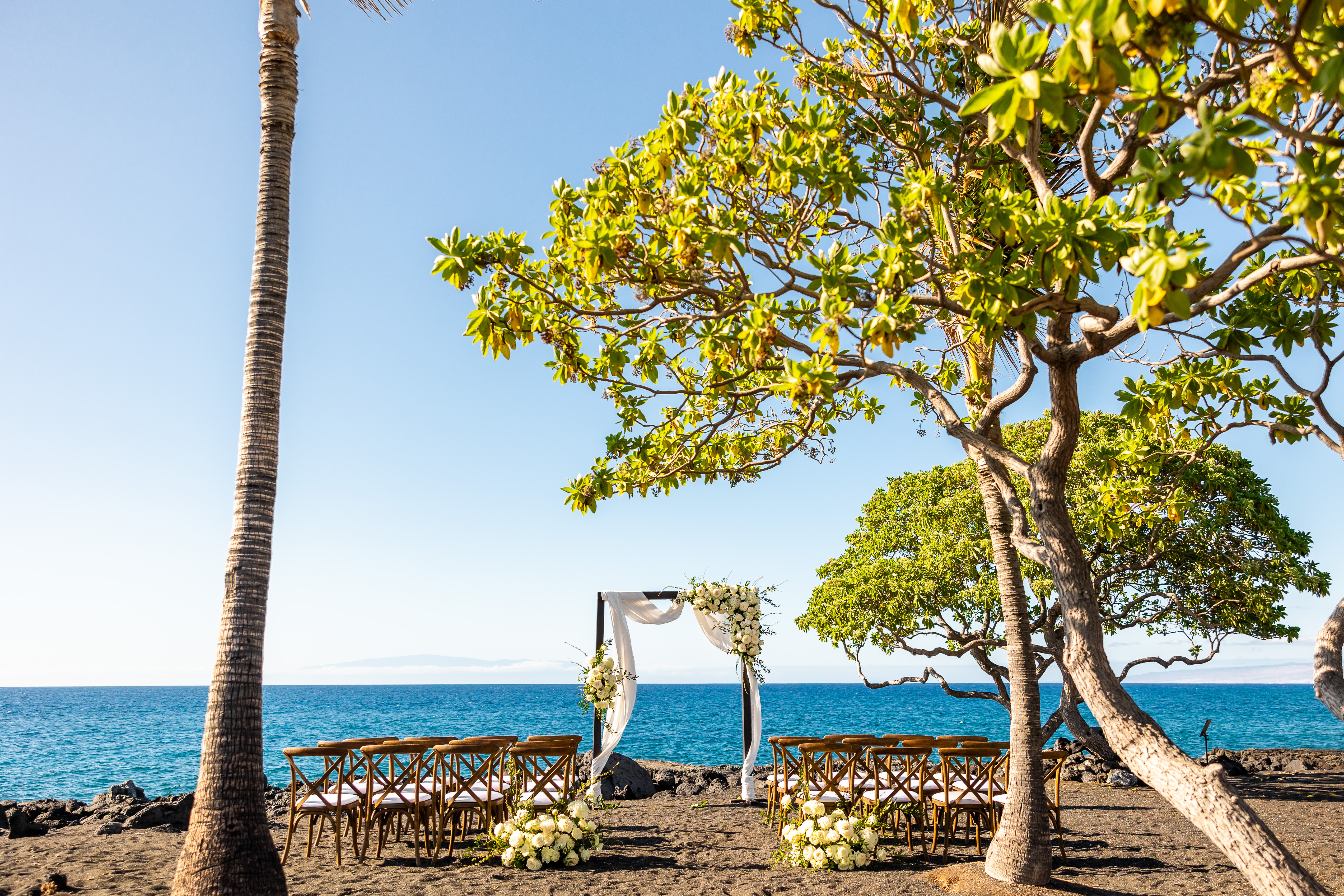 Beachfront wedding ceremony setup with floral arch, wooden chairs, and ocean backdrop.